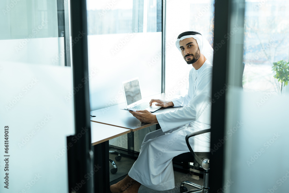 Fototapeta premium handsome man with dish dasha working in his business office of Dubai. Portraits of a successful businessman in traditional emirates white dress. Concept about middle eastern cultures.