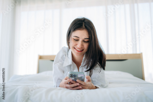 Young Woman Waking Up and play phone in Cozy Bedroom at Home. Beautiful Girl Smiling and Relaxing in Bed after Waking Up.
