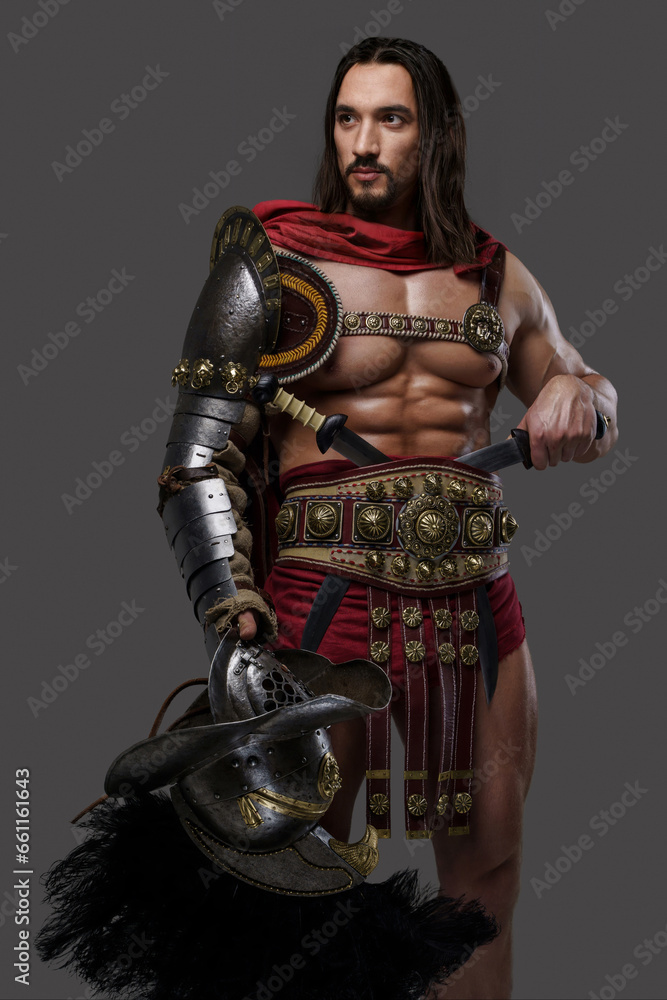 Muscular gladiator with a stylish beard and flowing hair dons intricate lightweight armor, holds a feathered helmet against a grey background