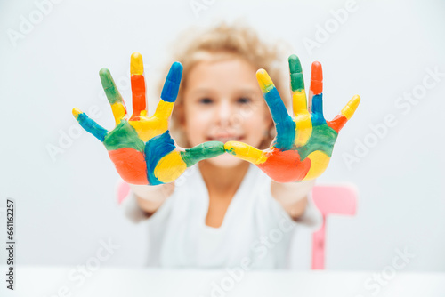 little blonde girl shows her hands painted with multi-colored paint on a white background.