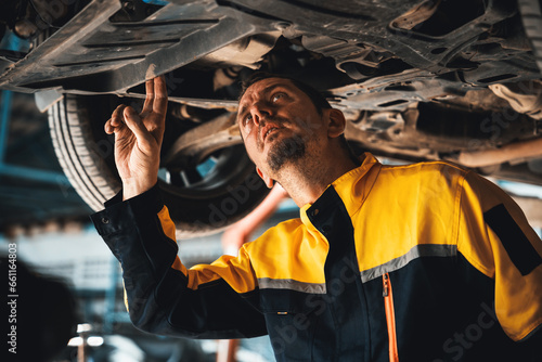 Vehicle mechanic conduct car inspection from beneath lifted vehicle. Automotive service technician in uniform carefully diagnosing and checking car's axles and undercarriage components. Oxus