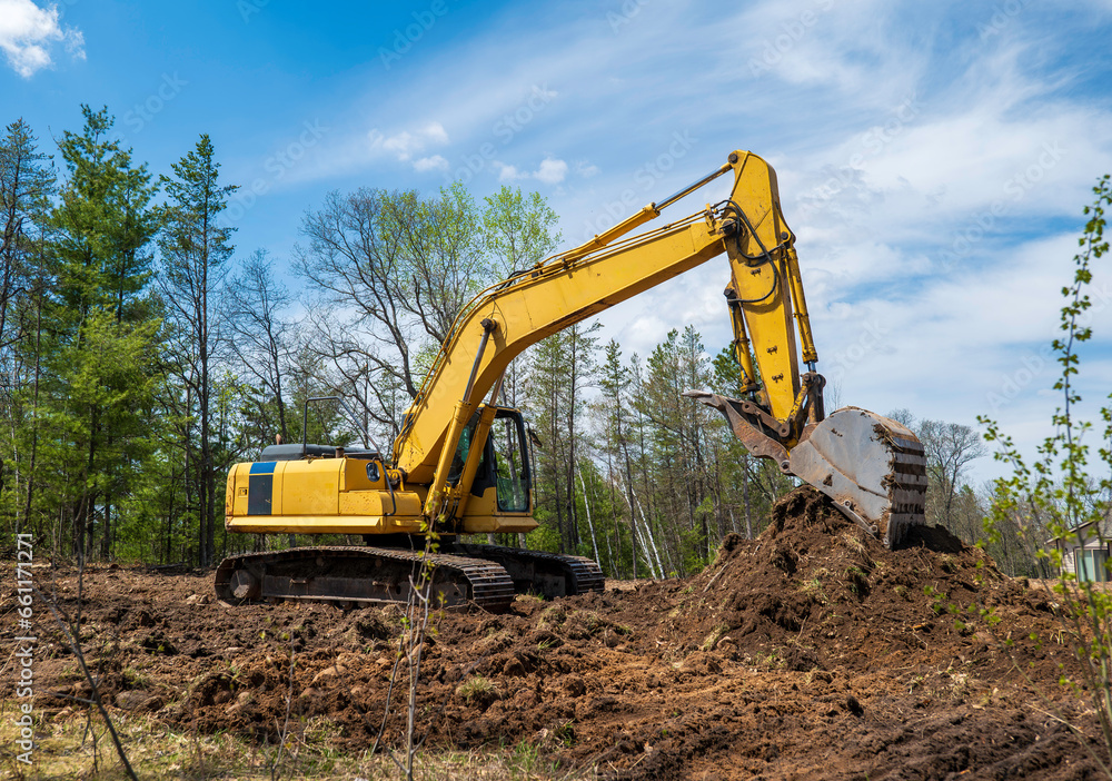 A yellow excavator at a future new home building site with its bucket resting on a pile of dirt. A yellow excavator at a future new home building site with its bucket resting on a pile of dirt.