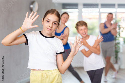 Joyful sister and brother practicing expressive dance movements against backdrop of mom and dad training together in wellness center
