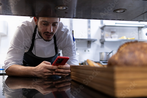 An arab chef is leaning against the kitchen while using his mobile phone.