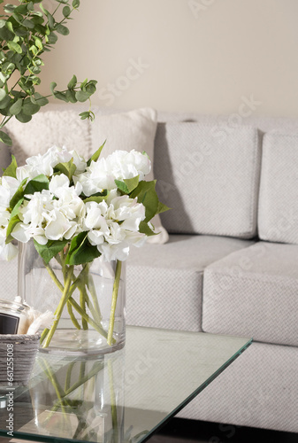 Close-Up of Delicate White Roses with Fresh Green Leaves in a Transparent Glass Vase, Set on a Glass Coffee Table, Glimpse of Textured Fabric Light Grey Sofa in the Backdrop, Soft Natural Light.