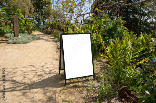 Blank white mockup template background texture of a sign board placed in an rural outdoor garden. Empty wedding or event information easel panel on footpath surrounded by green lush plants. photo
