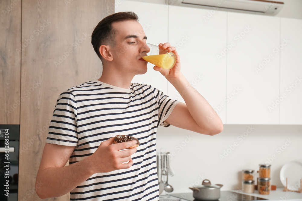 Fototapeta premium Young man with doughnut drinking orange juice in kitchen