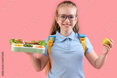 Happy little girl with backpack, lunchbox and apple on pink background