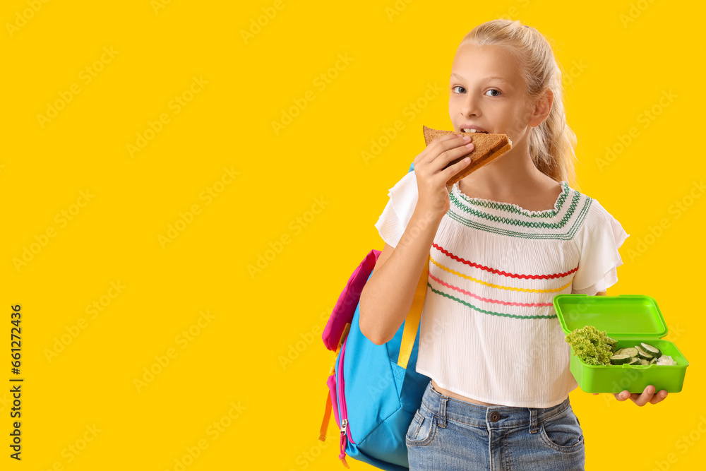 Happy girl with backpack and lunchbox eating sandwich on yellow background Happy girl with backpack and lunchbox eating sandwich on yellow background