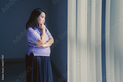 obese Asian woman looks pensive while standing by the window