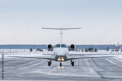 Front view of the modern white private jet taxiing on airport taxiway in winter