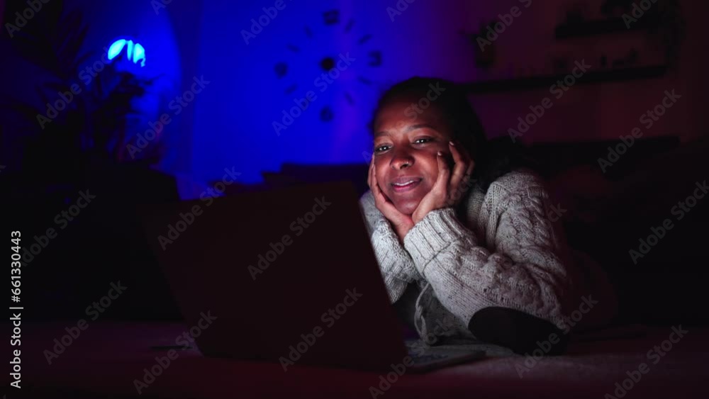 Smiling young black girl looking at laptop at home in evening. Latina woman lying on carpet cozy living room happy watching series or movie in dark. People enjoying domestic life and leisure time. Smiling young black girl looking at laptop at home in evening. Latina woman lying on carpet cozy living room happy watching series or movie in dark. People enjoying domestic life and leisure time.