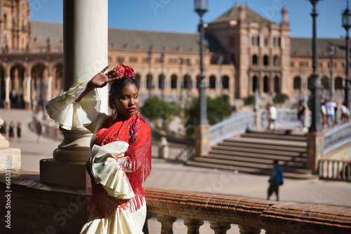 Young black and South American woman in a beige gypsy flamenco suit and red shawl, dancing in a beautiful square in the city of Seville in Spain. Concept dance, folklore, flamenco, art.