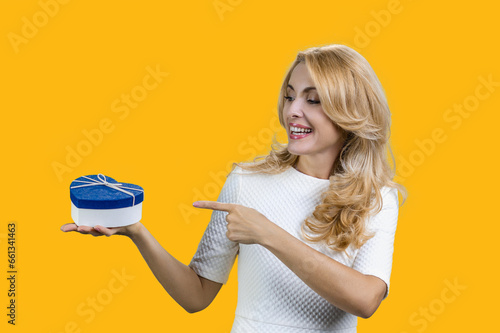 Happy blonde woman holding blue heart shape gift box and pointing with finger. Isolated on yellow.