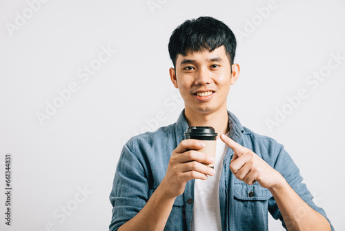 A hopeful young Asian man smiles, holding a coffee cup, and pointing with excitement. Studio portrait isolated on white with copy space. His positive expression is excellent.