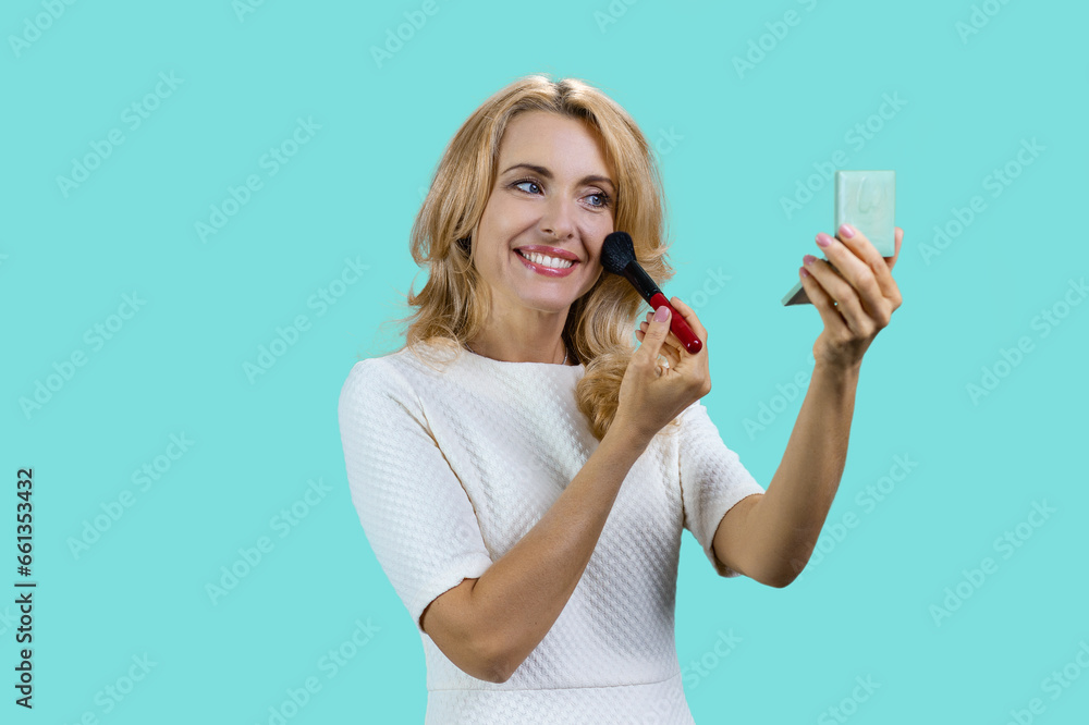 Happy blonde mature woman applying cosmetic by makeup brush. Isolated on turquoise.
