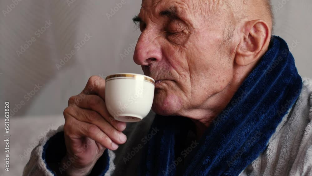 An elderly man in a bathrobe drinks tea and watches TV while sitting on the sofa at home. A hot drink in a cozy atmosphere. The concept of free time for a pensioner. An elderly man in a bathrobe drinks tea and watches TV while sitting on the sofa at home. A hot drink in a cozy atmosphere. The concept of free time for a pensioner.