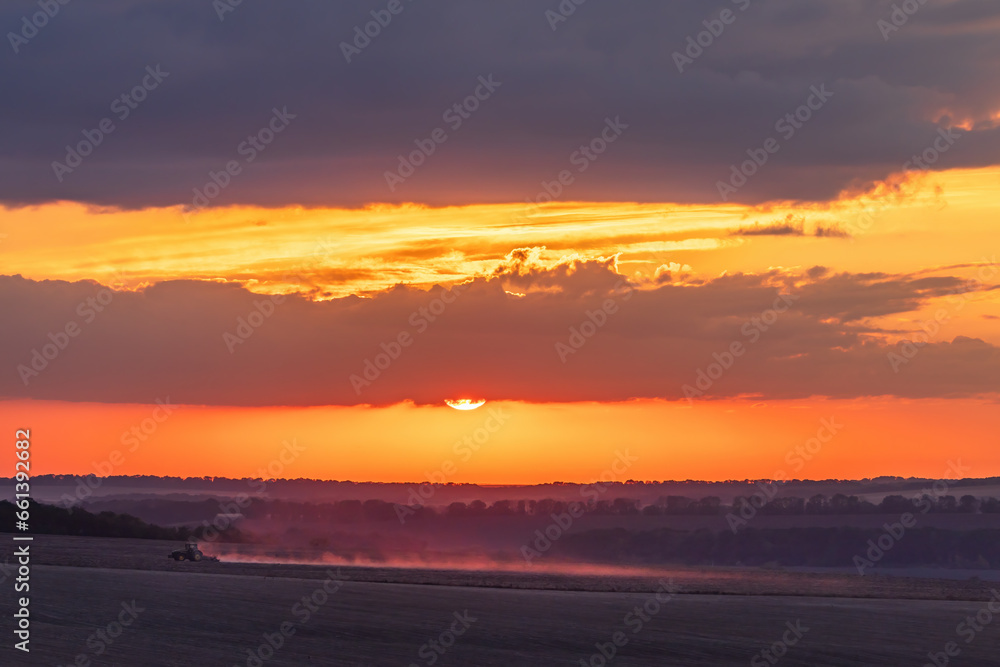 Tractor processes the fields during sunset and the dust glows in the dark under the sun and colorful sky