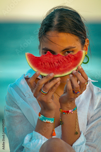 young woman modeling with sunglasses and a watermelon eating it wearing jewelry such as ring, bracelets and earrings. a white shirt on a beach day