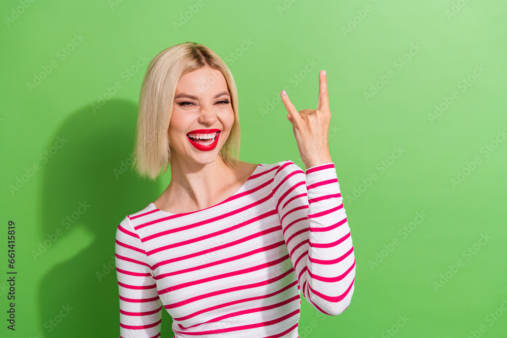 Photo of optimistic cool woman with bob hairdo dressed striped shirt fingers showing horns symbol isolated on green color background Photo of optimistic cool woman with bob hairdo dressed striped shirt fingers showing horns symbol isolated on green color background