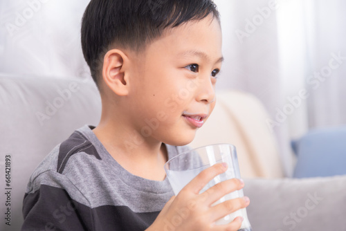 Close-up Asian preschool boy with milk mustache smile at unrecognizable mother sitting on sofa at home, young kid happy hold and drinking a clear glass full of dairy milk, healthy children's lifestyle