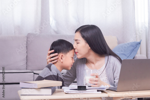 Happy single Asian mom drinks dairy milk from cute son hugging and kissing preschool boy bonding motherhood relationship, Asian freelancer woman remote working using modern technology laptop at home