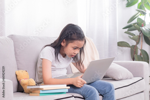 Asian sibling little girl playing game alone at home sitting on sofa using modern technology wireless laptop computer addiction, children studying homeschool learning typing keyboard listen to music