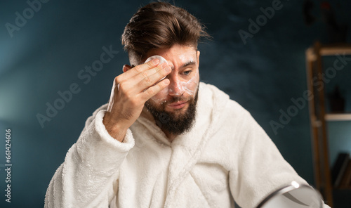 Attractive young man in white bathrobe sitting at vanity table with cosmetics at home, looking at mirror and using cotton pads for removing makeup © mtrlin