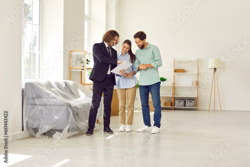 Young family buying new property. Happy married couple together with real estate agent sign contract in fully furnished white spacious Scandinavian living room interior in modern house or apartment