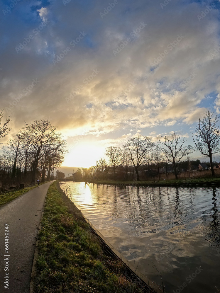 Nature's drama unfolds as the sun sets over a tranquil winter canal, painting the sky with breathtaking hues of orange, pink, and purple. Winter Canal Sunset. High quality photo