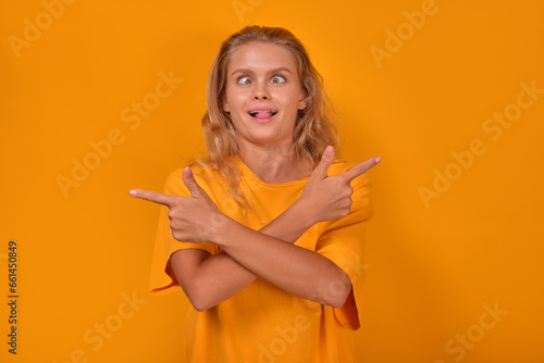 Young funny stupid Caucasian woman makes ridiculous grimace and points in different directions wanting to amuse children or draw attention to amazing promotional offer stands on yellow background.