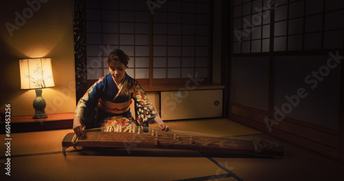 Talented Female Playing Koto Stringed Instrument in Traditional Dark Japanese Home with Lamp. Musician Wearing a Blue Kimono, Practicing to Play Historic Music Before a Performance