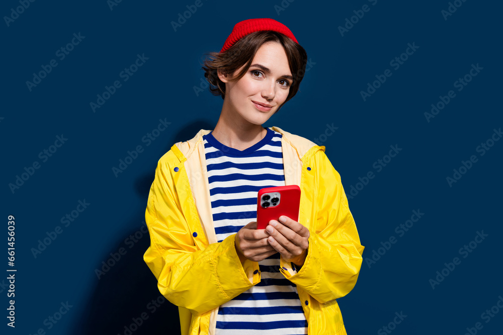 Photo of cute young hipster girl red beanie headwear browsing smartphone social media wear yellow raincoat isolated on blue color background Photo of cute young hipster girl red beanie headwear browsing smartphone social media wear yellow raincoat isolated on blue color background