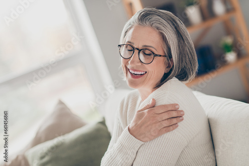 Photo of sweet adorable senior lady dressed white cardigan eyewear embracing herself closed eyes indoors house room