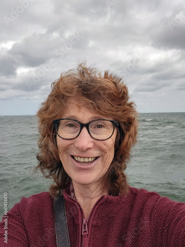 Mature woman takes a smiling selfie for social media at Mordialloc pier overlooking the sea. She is windswept and not wearing any make up. photo