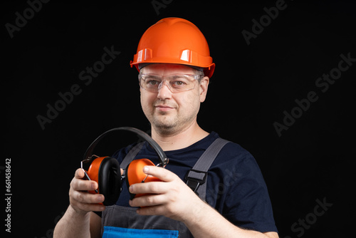 Builder in a protective helmet with noise-cancelling headphones in his hands on a black background, copy space for text photo