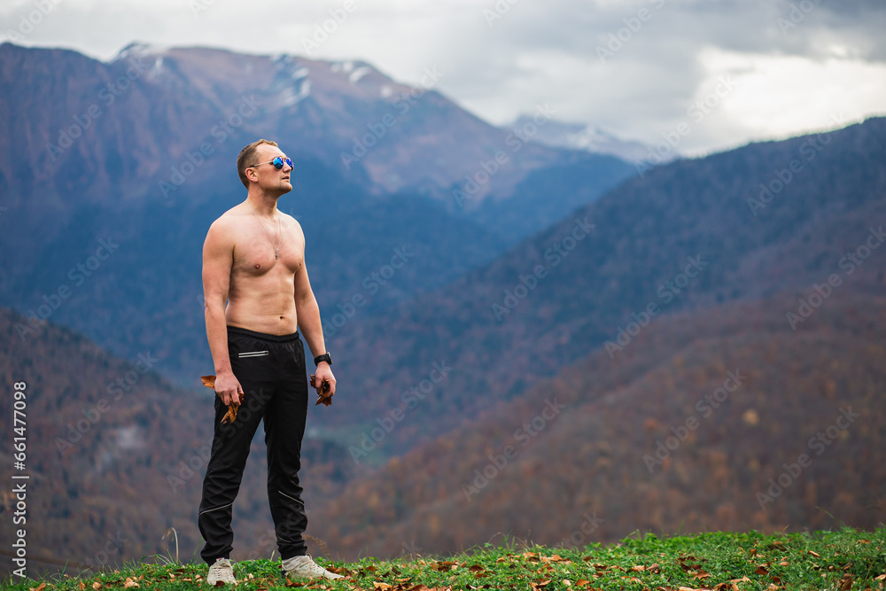 Young fitness trainer with naked torso doing aerobic exercises practicing his musculs, beautiful place on the top of the mountain with mountains landscape