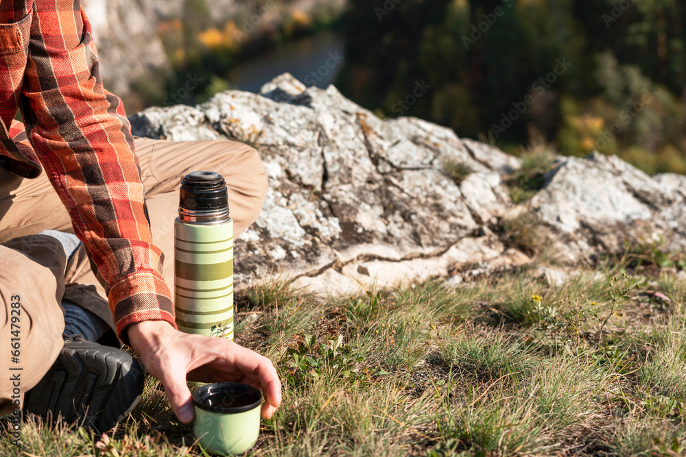 Fototapeta premium unrecognizable man tourist in red shirt drinking tea from thermos while resting on rocks by river in fall hiking and relaxing in nature