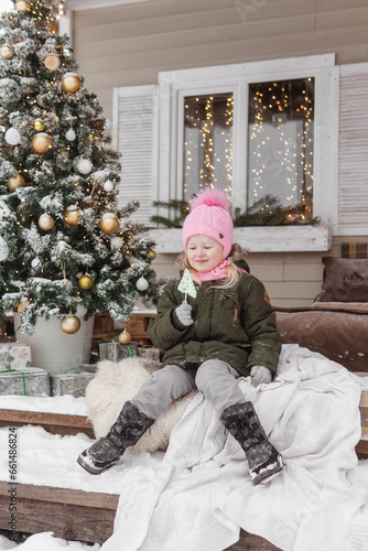 A little girl in winter clothes is sitting on the porch of a house decorated for the Christmas holidays. Christmas holidays for a child.