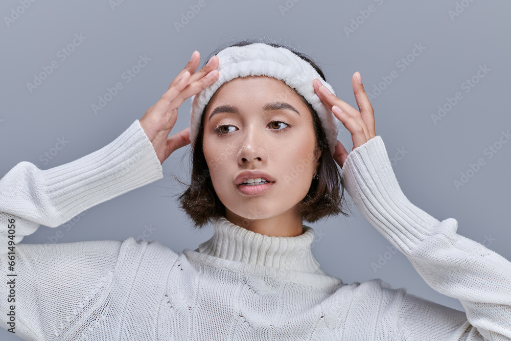 Obraz premium stylish asian woman in knitted sweater adjusting warm headband on grey, winter fashion photography