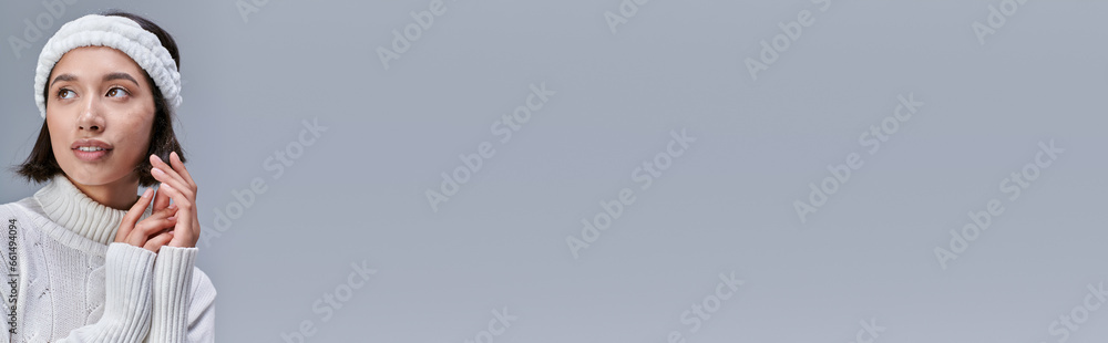 fashionable and dreamy asian model in white sweater and warm headband looking away on grey, banner fashionable and dreamy asian model in white sweater and warm headband looking away on grey, banner
