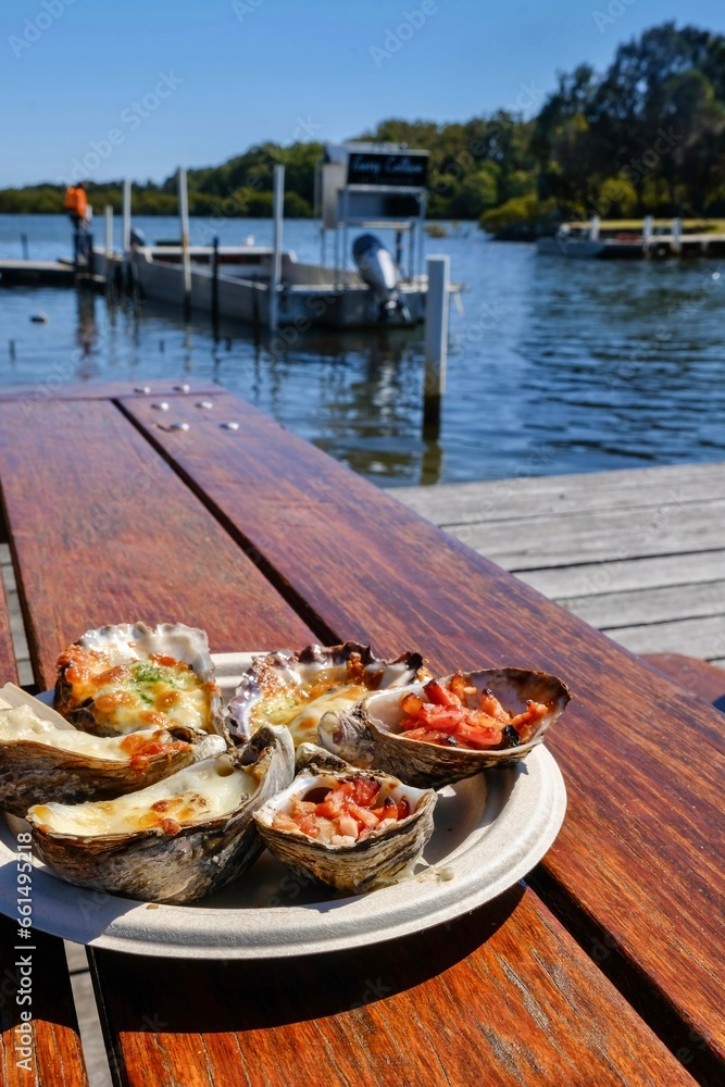 A plate of grilled oysters with assorted toppings (garlic cheese, mornay, bacon) at Jim Wilds Oyster Shack, Greenwell Point, NSW with view of Crookhaven River 