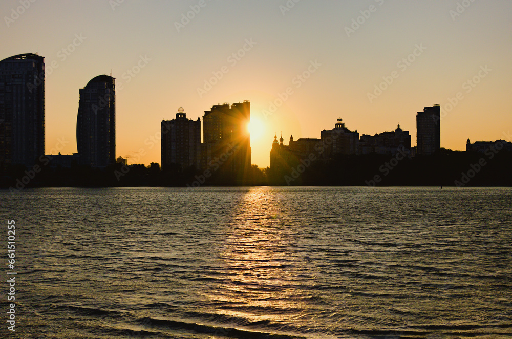 Scenic sunset landscape view of black silhouettes of high-rise buildings along the Dnipro River. Obolon neighborhood in Kyiv. Ukraine. Sun hides behind the skyscraper