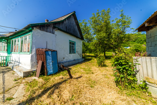 a dilapidated one-story rural house. Green lawn and trees. Whitewashed walls, green window frames.