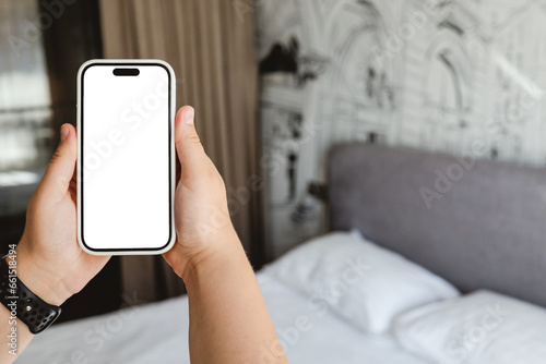 Woman hand holding a modern frameless smartphone with blank white screen on background of a modern bedroom interior