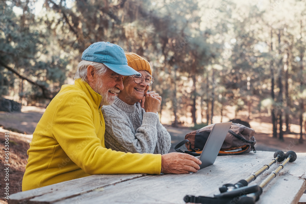 Head shot portrait close up of cute couple of old middle age people using computer pc outdoors sitting at a wooden table in the forest of mountain in nature with trees around them.. Head shot portrait close up of cute couple of old middle age people using computer pc outdoors sitting at a wooden table in the forest of mountain in nature with trees around them..