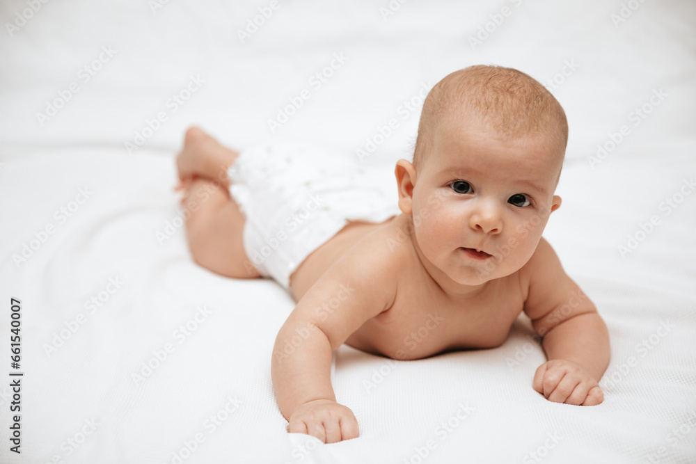 A newborn baby in a diaper is lying on stomach on a white background and looking at the camera. 