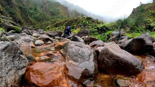 A man walking over the rocks inside a river in a rainy day with a raincoast, surrounded of green grass, moss, in a gorge in Azores, in the trail Valle das Lombadas photo