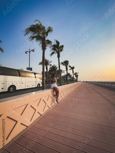 Dubai, UAE. September 27, 2023: a cute girl sits on a fence in shorts and a white tourist bus passes behind her