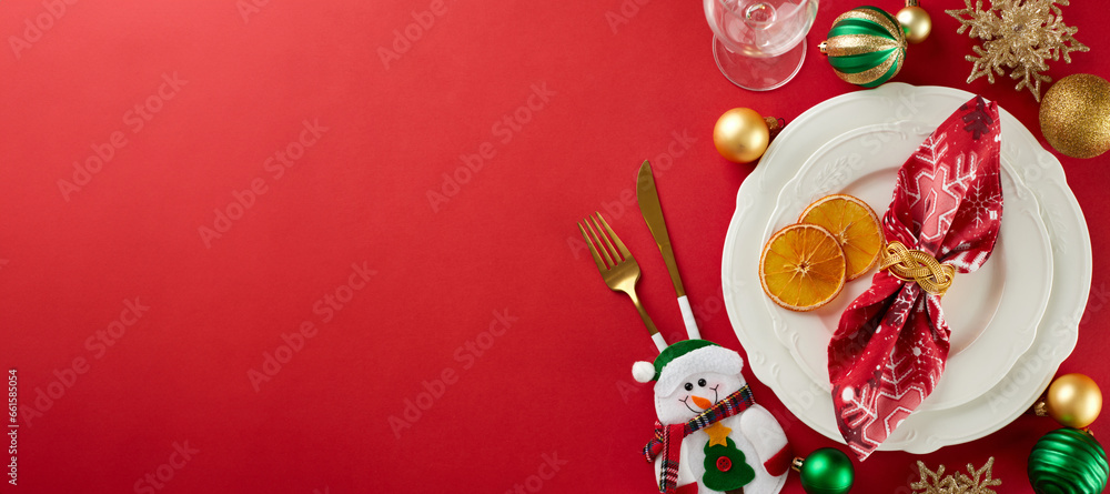 Elevate your Christmas dining experience. Top view of plates, cutlery, glass, snowflakes, xmas balls, snowman, dry orange slices on red background with advert space